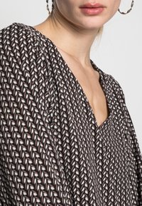 Blouse marron à motifs avec un décolleté en V profond, manches longues et tissu texturé doté de motifs en zigzag blancs et crème.