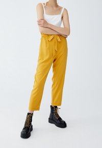 Gula skräddarsydda byxor med bälte i midjan och smalnande ben. Matchas med svarta snörkängor med guldfärgade ögla.