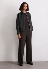 Donkergrijze jas met een ritssluiting aan de voorkant en opstaande kraag, gecombineerd met bruine broek. Glad textuur en ontspannen pasvorm, minimale details.