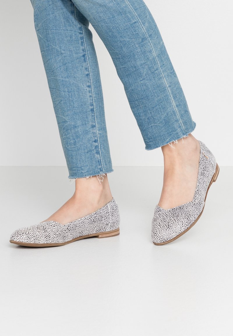 Personne portant un jean bleu court avec des ourlets effilochés et des chaussures plates blanches avec un motif noir moucheté sur un sol blanc.