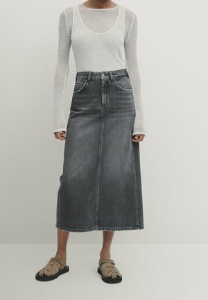 Personne portant un haut blanc transparent à manches longues, une jupe midi en denim gris foncé taille haute et des sandales beiges à boucle, debout devant un fond uni.