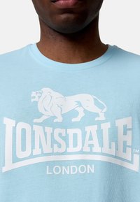 Camiseta de algodón azul cielo con un gran gráfico de un león blanco y el texto "LONSDALE LONDON". Diseño clásico de cuello redondo, textura suave.