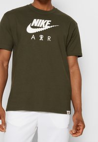 Olivově zelené bavlněné tričko s bílým logem Nike a textem "AIR" na přední straně. Krátké rukávy, kulatý výstřih, pohodlný střih.