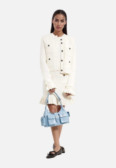 Veste texturée crème avec jupe assortie ; présente des poignets et un ourlet ruffles. Tient un sac à main léger bleu brillant avec poches avant. Chaussures plates noires.