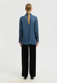 Blusa de seda azul con mangas largas, puños abotonados y corte recto, combinada con pantalones negros de pierna ancha, mostrada desde la parte posterior.