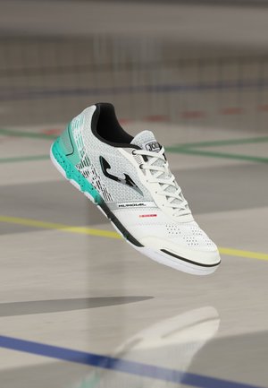 Weißer und türkisfarbener Hallenfußballschuh mit schwarzem Logo, Schnürsenkeln und Sohlen-Details, in der Luft schwebend auf einem Hallenboden mit bunten Linien gezeigt.