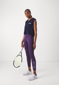 Marineblå ermeløs topp med hvit "Lacoste" logo, kombinert med lilla leggings med lilla "Lacoste" tekst, og hvite joggesko.