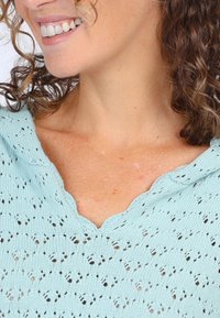 Helles blaues, gestricktes Oberteil mit einem gewellten Ausschnitt und einem Lochmuster. Lockige Haare teilweise sichtbar, zeigen Hautton und Details des Ausschnitts.