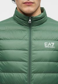 Grön vadderad jacka med hög krage och dragkedja framtill. Den har en quiltad design och visar en vit EA7-logotyp på bröstet.