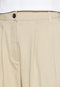Beige geplooide broek met een gestructureerde afwerking, voorzien van een voordoek en een zwarte knoopsluiting bij de tailleband.
