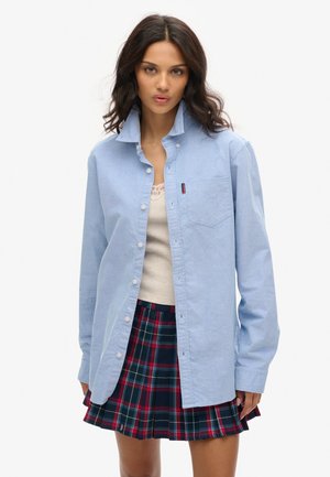 Lyseblå skjorte med knapper, krave og brystlomme, kombineret med et plisseret ternet skørt med mørkegrønne og røde detaljer.