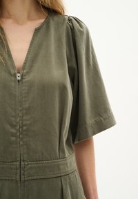 Salopetă de culoare verde măslinie, cu mâneci scurte, cu fermoar frontal, detalii pliate pe umeri și talie ajustată, confecționată dintr-un material texturat din bumbac.