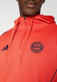 Sweat à capuche rouge avec accents noirs, arborant le logo du FC Bayern München et un col zippé. Tissu lisse avec une coupe structurée.