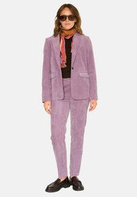 Costume en velours lilas avec une veste sur mesure et un pantalon ajusté. Associé à un haut noir à côtes, une écharpe colorée et des mocassins foncés.