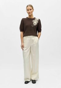Femme debout portant un pull marron à manches courtes avec une broderie florale beige et un pantalon crème à jambes larges et taille haute, accompagné de chaussures noires.