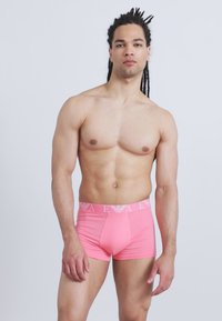 Μυώδης άνδρας με μακριά dreadlocks φοράει φωτεινά ροζ μπόξερ και στέκεται μπροστά σε απλό ανοιχτό γκρι φόντο.