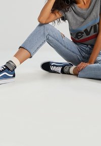 Persona vistiendo jeans rasgados de color azul claro, calcetines a rayas, zapatillas azules y negras, y una camiseta gris de Levi's sentada en el suelo.