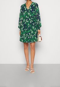 Sukienka w zielony liść z dekoltem w kształcie V, długimi rękawami i falbaniastym dołem. Połączona z białymi sandałami na paski i małą torebką.