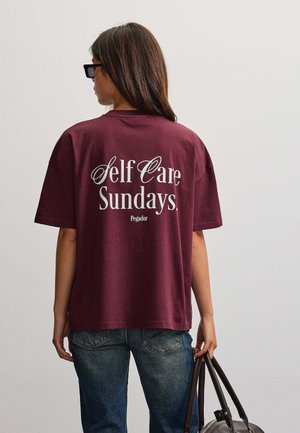 Frau trägt ein weinrotes Hemd mit dem Text "Self Care Sundays", blaue Jeans, hält eine dunkle Handtasche und steht mit dem Rücken zu einem einfarbigen Hintergrund.