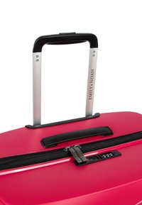 Červený kufr s tvrdou skořápkou a hliníkovou teleskopickou rukojetí, černý zip a kombinovaný zámek. Vyznačuje se texturovaným povrchem a detailem s logem.