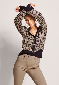 Strickjacke mit geometrischem Muster in Beige und Dunkelblau, mit gerippten Bündchen und Knopfleiste. Kombiniert mit beigen Hosen.