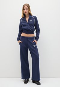 Giacca cropped blu navy con zip e bordi bianchi, abbinata a pantaloni della tuta a gamba larga con un design ricamato. Tessuto morbido e liscio.