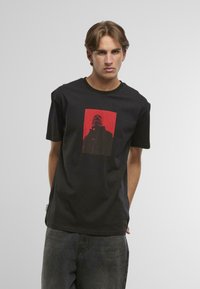 Mladý muž s krátkymi hnedými vlasmi, ktorý má na sebe čierne tričko s červeným a čiernym grafickým motívom osoby, ktorá sa pozerá hore.