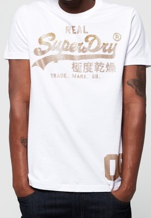 Biała bawełniana koszulka z teksturowanym, złoto-czarnym nadrukiem logo "SuperDry" oraz małą grafiką na lewym brzegu. Krótkie rękawy, standardowy krój.