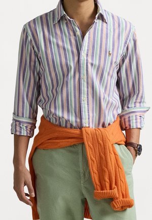 Mand iført pastelstribet skjorte med knapper, orange sweater bundet omkring taljen, lysegrønne bukser og læderur på håndleddet.