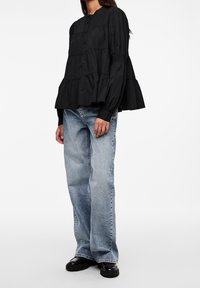 Blusa nera con maniche a sbuffo, design a strati e chiusura con bottoni; abbinata a jeans azzurri larghi e scarpe nere.