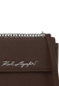 Borsa a tracolla in pelle marrone con lavorazione testurizzata, dotata di una tracolla in catena argentata e dettagli con logo distintivo. Presenta una chiusura a flap e un design strutturato.