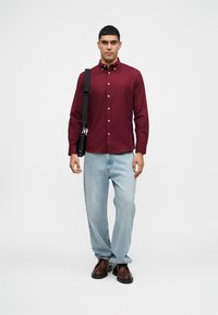 Burgundski košulja na gumbi, svijetloplave traperice, smeđe cipele, crna torba preko ramena, jednostavna pozadina. Košulja ima gumbe i ovratnik.