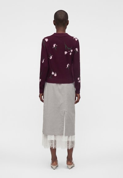 Personne de dos, portant un pull bordeaux à motifs floraux, une jupe grise fendue à l'arrière, un ourlet en dentelle blanche et des chaussures beiges sur fond uni.
