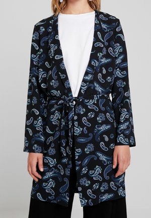 Kvinde iført en sort langærmet blazer med blå paisley- og blomsterprint, bundet i taljen, over en ensfarvet hvid top.
