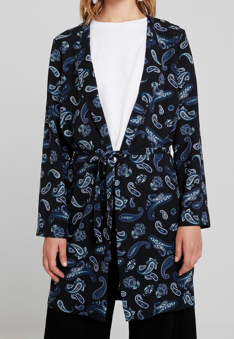 Femme portant un blazer noir à manches longues avec des motifs paisley et floraux bleus, noué à la taille, sur un haut blanc uni.