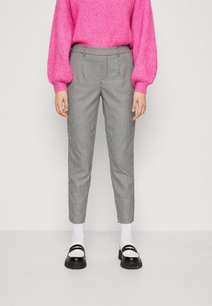 Personne portant un pull rose vif, un pantalon slim à carreaux gris, des chaussettes blanches et des mocassins noir et blanc, se tenant devant un fond uni.