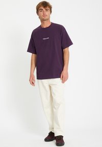 Lilla bomulds-t-shirt med korte ærmer og hvid "VOLCOM" logo, parret med cremefarvede bukser og sorte sko med mønstrede snørebånd.
