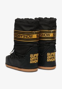 Botas de nieve negras con parte superior acolchada, que presentan una banda a rayas dorada y negra y un logo en relieve. Suela de goma con un acabado texturizado.
