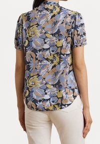 Blusa floreale di colore blu scuro, con una varietà di fiori in delicati toni di blu, beige e giallo. Maniche a palloncino corte e orlo arrotondato.