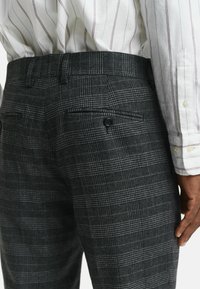 Man in donkergrijze geruite broek en een wit overhemd met lange mouwen met dunne verticale strepen, staande met zijn rechterhand zichtbaar.