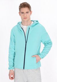 Felpa leggera con zip di un vivace verde menta, con dettagli in blu navy, dotata di cappuccio, tasche laterali e una texture liscia.