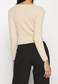 Kvinna klädd i en ribbad, långärmad, beige cropped tröja som är kombinerad med högmidjade svarta byxor, sedd bakifrån.