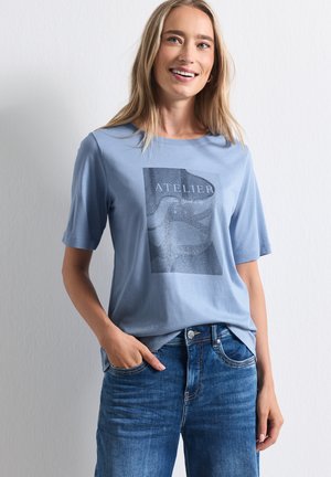 Donna sorridente con capelli biondi lunghi che indossa una maglietta azzurro chiaro con grafica e jeans blu, in piedi con una mano in tasca.
