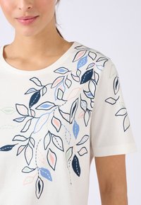 Mujer con una camisa blanca de manga corta con un patrón multicolor de hojas botánicas en azul, rosa, marino y verde en el hombro y el pecho.