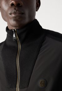 Jachetă neagră cu fermoar, având un guler înalt, material texturat, panouri netede și un fermoar auriu. Include un patch cu logo negru pe piept.