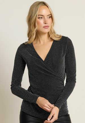 Blond kvinde iført en langærmet sort shimmer-wrap top, ser til siden med hænderne foldet om livet mod en ensfarvet baggrund.