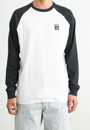 Mężczyzna ubrany w białą koszulę z długim rękawem, z czarnymi kropkowanymi rękawami i logo Adidas na piersi, w połączeniu z jasnoszarymi spodniami.