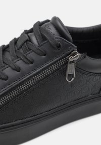 Baskets noires avec un motif texturé sur le côté, lacets noirs et une fermeture éclair métallique avec le logo Calvin Klein sur le côté.
