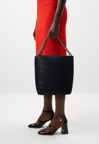 Sac fourre-tout en cuir noir avec une forme structurée, une anse en chaîne et un accent logo. Les détails comprennent une texture lisse et un éclat subtil.
