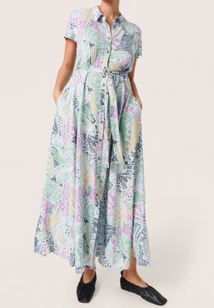 Kvinde iført en pastelblomstret maxi-kjole med knapper foran, korte ærmer og bindebånd i taljen, kombineret med sorte flade sko.